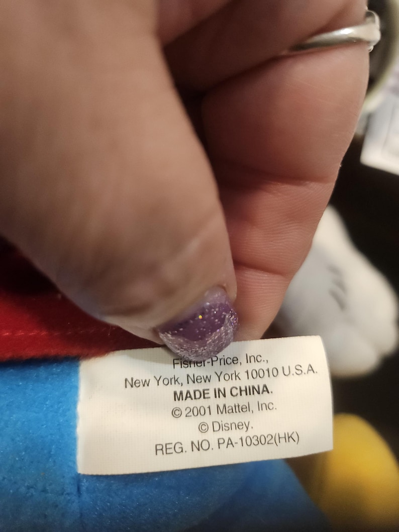 May include: A close-up of a white tag with black text from a Fisher-Price toy. The tag includes the company's address, the phrase "MADE IN CHINA," and copyright information for Mattel and Disney. The tag is held by a hand.
