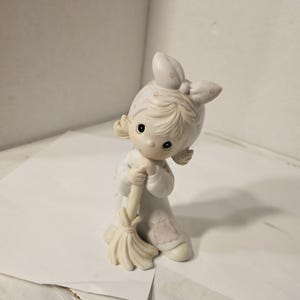 May include: A ceramic figurine of a young girl in a nightgown and bonnet, holding a broom. The figurine is primarily off-white with subtle pink accents on the patch of the nightgown. The girl has dark eyes and blonde hair.