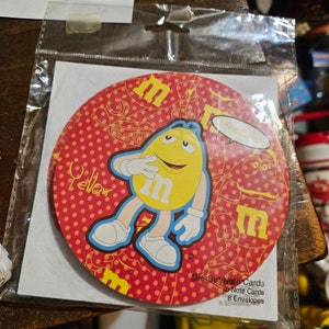 May include: A red and yellow round card featuring a yellow M&M character wearing white gloves and a red and white hat. The card has white polka dots and the text "Yellow" and "Life is Sweet!" printed on it. The card is part of a set of Die Cut Note Cards.
