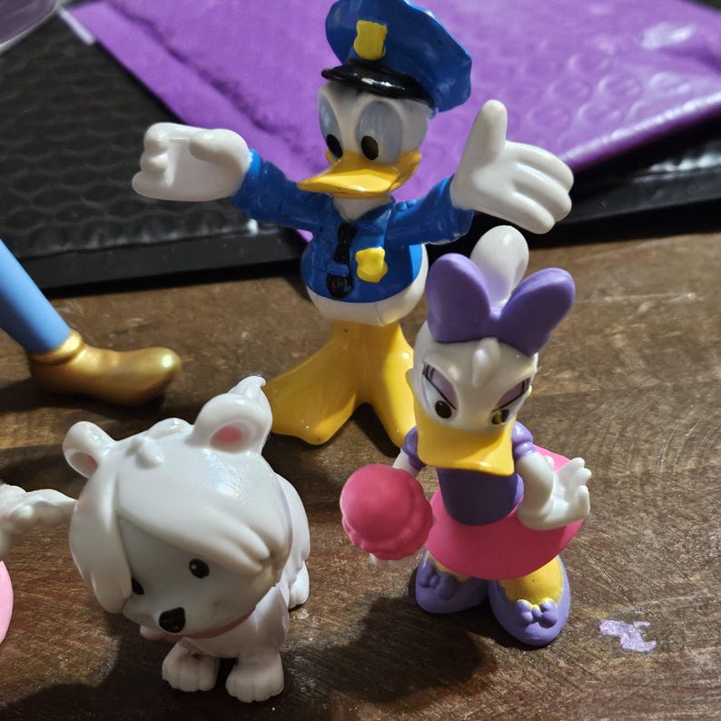 Może przedstawiać: Kolekcja figurek postaci Disneya, w tym Kaczor Donald w mundurze policjanta, Daisy Duck i biała figurka przypominająca szczeniaka. Figurki są w jasnych kolorach i wykonane z tworzywa sztucznego, z detalami.