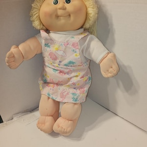 以下が含まれることがあります： 丸い顔、ブロンドの糸の髪、緑の目をしたヴィンテージ人形。人形は、動物と雲の模様のピンク色のオーバーオールドレスの下に白いシャツを着ています。人形は柔らかいぬいぐるみのような腕と脚を持っています。