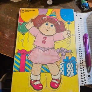 May include: A 9-piece jigsaw puzzle featuring a cartoon girl with a pink dress and a birthday hat. The puzzle pieces are on a yellow background with colourful balloons and presents.