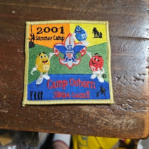 May include: A yellow, green, blue, and red embroidered patch with the text "2001 Summer Camp Camp Osborn SWGA Council" and a scout emblem surrounded by M&M characters.