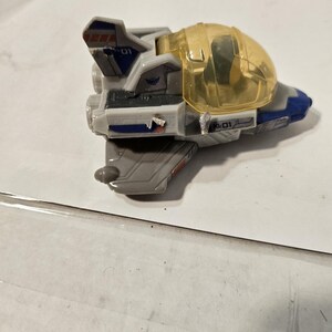 May include: A gray and blue toy spaceship with a clear yellow cockpit. The toy has the number "01" printed on the side and top. The spaceship has red and blue accents.
