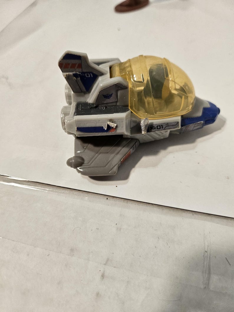May include: A gray and blue toy spaceship with a clear yellow cockpit. The toy has the number "01" printed on the side and top. The spaceship has red and blue accents.