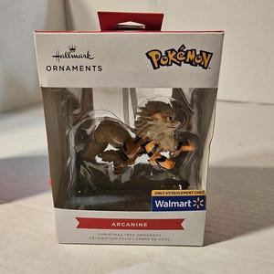 May include: A Hallmark Christmas tree ornament featuring the Pokémon character Arcanine. The figure is brown and orange, displayed in a clear plastic case within a branded box. The box includes the Walmart logo and the text "Christmas Tree Ornament".