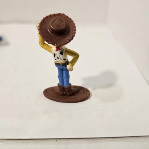 May include: A small figurine of a cowboy character, wearing a brown hat, yellow shirt, blue jeans, and brown boots. The cowboy has a white vest with black spots and a red neckerchief. The figurine is standing on a brown oval base.
