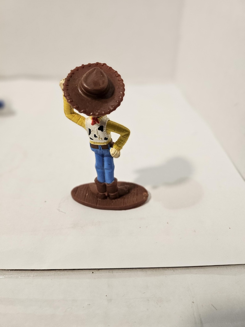 May include: A small figurine of a cowboy character, wearing a brown hat, yellow shirt, blue jeans, and brown boots. The cowboy has a white vest with black spots and a red neckerchief. The figurine is standing on a brown oval base.