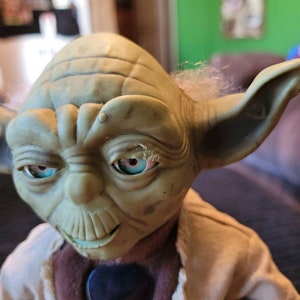K&ouml;nnte beinhalten: Eine gr&uuml;ne Yoda-Puppe mit einem braunen Gewand und einem schwarzen Gitarrenplektrum. Die Puppe hat einen wei&szlig;en Bart und blaue Augen.