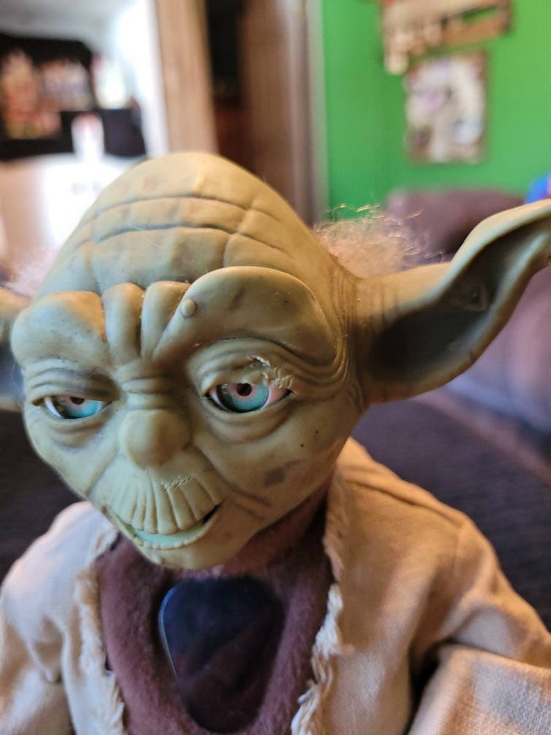 K&ouml;nnte beinhalten: Eine gr&uuml;ne Yoda-Puppe mit einem braunen Gewand und einem schwarzen Gitarrenplektrum. Die Puppe hat einen wei&szlig;en Bart und blaue Augen.