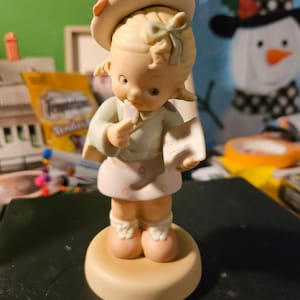 以下が含まれることがあります： ピンクと白のドレスとピンクの帽子をかぶった女の子の磁器製フィギュア。白い花と「あなたへ」と書かれたメモを持っています。
