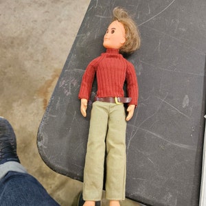 May include: A red and brown doll with brown hair wearing a red sweater and brown pants with a brown belt.