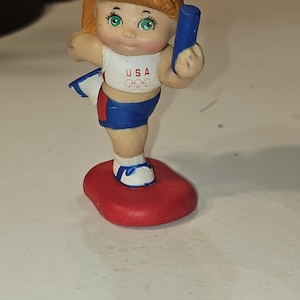 May include: A figurine of a young athlete in a USA uniform, holding a blue baton. The figure has blonde hair and green eyes, standing on a red base. The uniform includes a white top with "USA" and Olympic rings, and blue shorts.