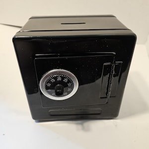 Puede incluir: Una caja fuerte negra y rectangular con un dial de combinación y una pequeña ranura en la parte superior. La caja fuerte tiene una puerta con bisagras y una superficie brillante y reflectante. El dial de combinación tiene números del 0 al 40.