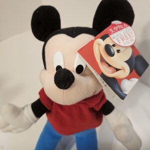 May include: A Mickey Mouse soft toy with black ears, a black nose, and white gloves. Mickey is dressed in a red top, blue trousers, and yellow shoes. A red tag with Mickey's image and text is attached.