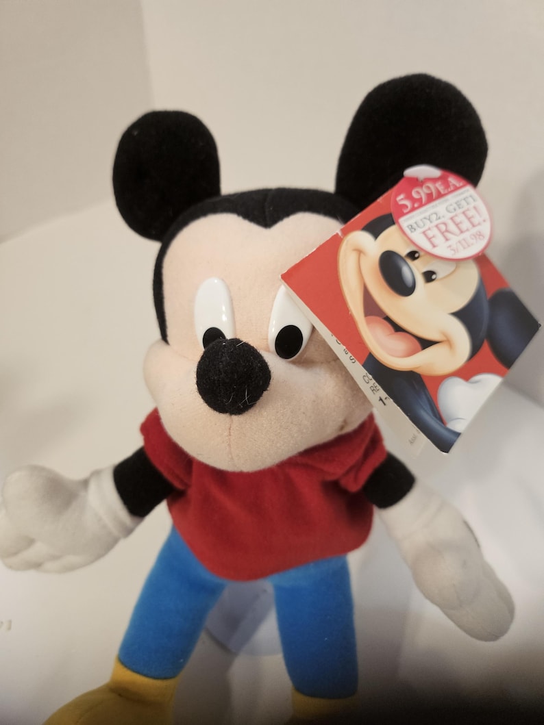 May include: A Mickey Mouse soft toy with black ears, a black nose, and white gloves. Mickey is dressed in a red top, blue trousers, and yellow shoes. A red tag with Mickey's image and text is attached.