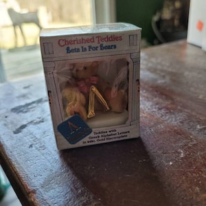 May include: A small teddy bear holding a gold letter A. The teddy bear is wearing a pink bow. The box reads "Cherished Teddies Beta Is For Bears" and "Teddies with Greek Alphabet Letters in 24kt. Gold Electroplate".