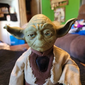 K&ouml;nnte beinhalten: Eine gr&uuml;ne Yoda-Puppe, die eine braune Robe mit einem Fransenkragen und einem braunen G&uuml;rtel tr&auml;gt. Die Puppe hat einen gro&szlig;en Kopf mit gro&szlig;en Ohren und einem breiten Mund. Die Augen der Puppe sind blau und die Puppe schaut direkt in die Kamera.