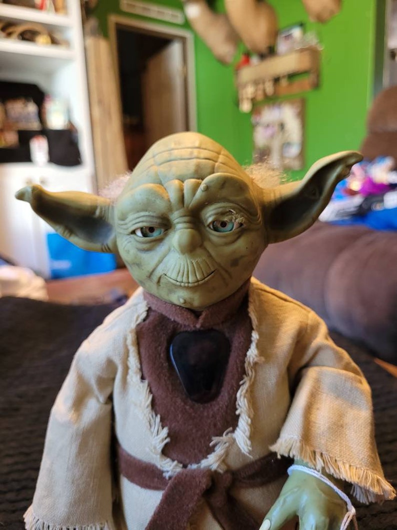 K&ouml;nnte beinhalten: Eine gr&uuml;ne Yoda-Puppe, die eine braune Robe mit einem Fransenkragen und einem braunen G&uuml;rtel tr&auml;gt. Die Puppe hat einen gro&szlig;en Kopf mit gro&szlig;en Ohren und einem breiten Mund. Die Augen der Puppe sind blau und die Puppe schaut direkt in die Kamera.