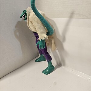 May include: A green and purple action figure of a lizard-like creature with a long, segmented teal tail. The figure wears a tattered, off-white cloth and purple pants. The creature has a detailed, reptilian head.