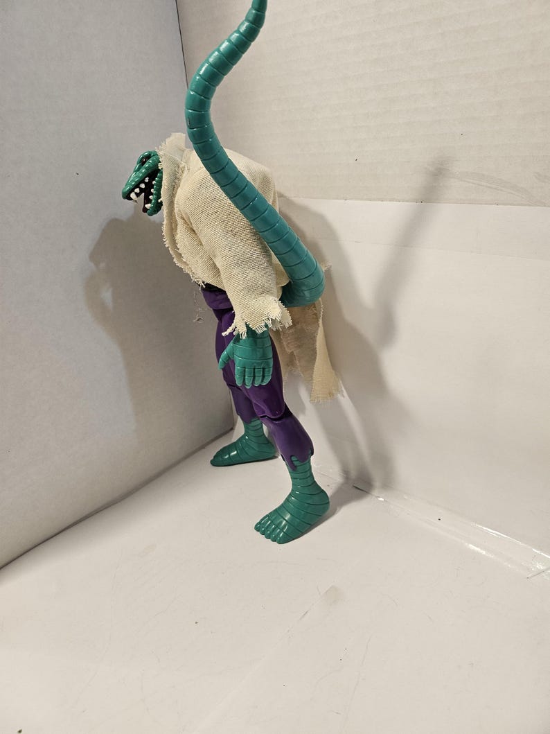 May include: A green and purple action figure of a lizard-like creature with a long, segmented teal tail. The figure wears a tattered, off-white cloth and purple pants. The creature has a detailed, reptilian head.
