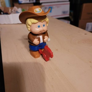 May include: A wind-up toy of a boy wearing a brown cowboy hat with a horseshoe design, a brown jacket, blue jeans, and brown boots. The boy is riding a red toy horse.