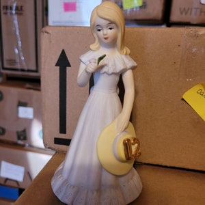 May include: A porcelain figurine of a girl in a white dress holding a yellow hat with the number 12 on it. She is holding a small pink flower in her hand.
