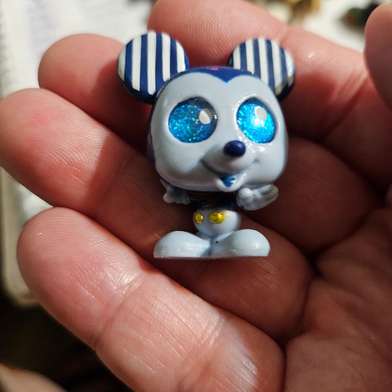 Peut inclure: Une petite figurine en plastique bleue de Mickey Mouse avec des yeux bleus scintillants. La figurine porte un chapeau ray&eacute; et un short rouge avec des boutons jaunes.