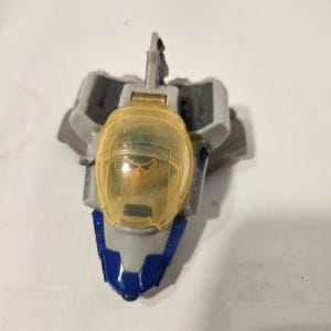 May include: A toy spaceship with a clear yellow cockpit and a blue and gray body. The cockpit contains a small figure. The spaceship has gray wings and a blue nose. The toy is designed for children.