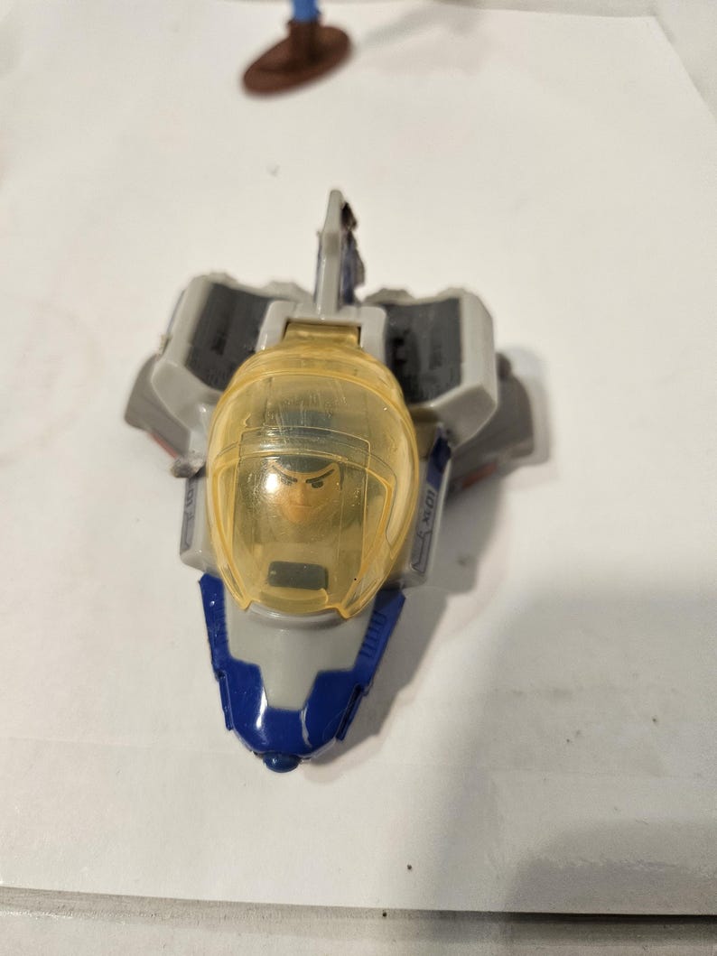 May include: A toy spaceship with a clear yellow cockpit and a blue and gray body. The cockpit contains a small figure. The spaceship has gray wings and a blue nose. The toy is designed for children.