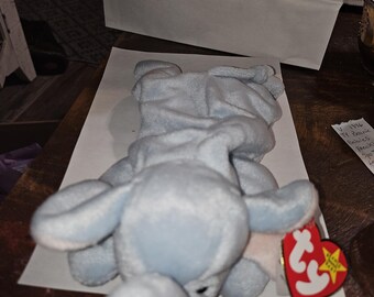 May include: A light blue plush elephant toy with a Ty tag attached. The elephant is lying on its side with its trunk curled up.