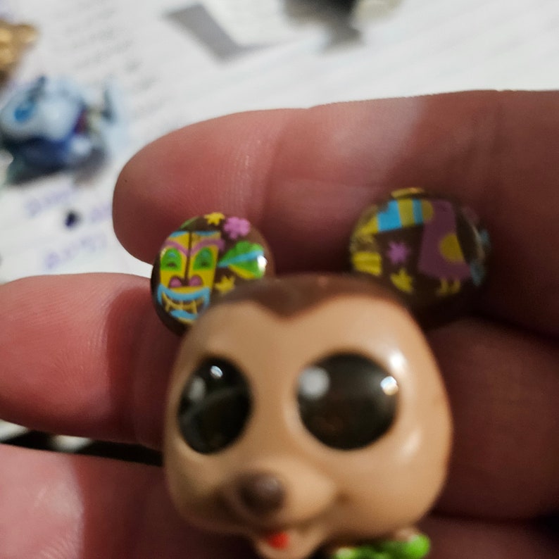Peut inclure: Un jouet en peluche brun et beige avec de grands yeux et un nez brun. Le jouet a deux oreilles avec des motifs color&eacute;s, l'une avec un masque tiki et l'autre avec un oiseau tropical et des fleurs.