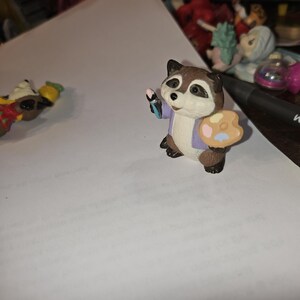 Puede incluir: Una pequeña figura de un mapache vestido con una chaqueta morada y una camisa blanca, sosteniendo un pincel y una paleta. El mapache tiene una cara marrón y blanca y ojos negros. La paleta tiene pintura rosa, amarilla y azul.