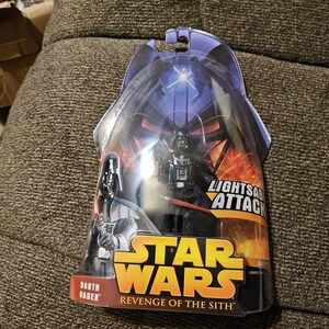 May include: Darth Vader action figure from Star Wars: Revenge of the Sith. The figure is packaged in a plastic blister pack with a blue and red background. The figure is holding a lightsaber and has a Lightsaber Attack feature.