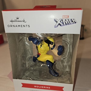 May include: A Hallmark Christmas tree ornament featuring Wolverine from Marvel's X-Men. The figurine is in a dynamic pose, wearing a yellow and blue costume. The packaging includes the Hallmark Ornaments logo and the Marvel X-Men logo.