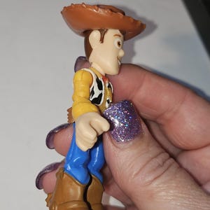 Op de afbeelding: Een kleine plastic speelgoedfiguur van een cowboy-personage, met een bruine hoed, geel overhemd, zwart-wit vest, blauwe broek en bruine laarzen. De figuur wordt in een hand gehouden.