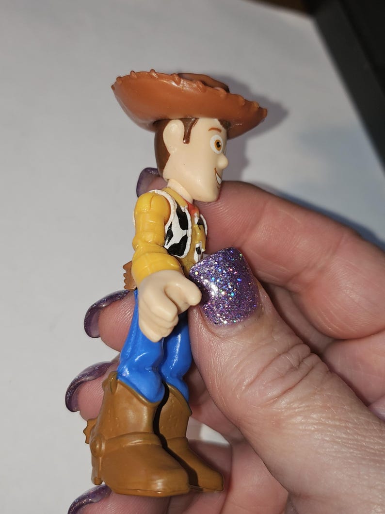 Op de afbeelding: Een kleine plastic speelgoedfiguur van een cowboy-personage, met een bruine hoed, geel overhemd, zwart-wit vest, blauwe broek en bruine laarzen. De figuur wordt in een hand gehouden.
