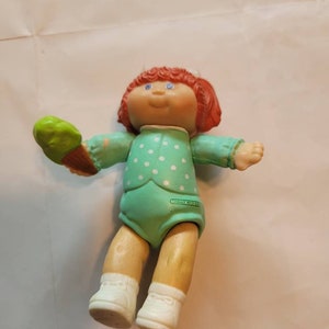 Op de afbeelding: Een Cabbage Patch Kid pop met rood haar, die een groen-wit geblokt jurkje draagt en een groene ijshoorn vasthoudt. De pop heeft blauwe ogen en een glimlachend gezicht.