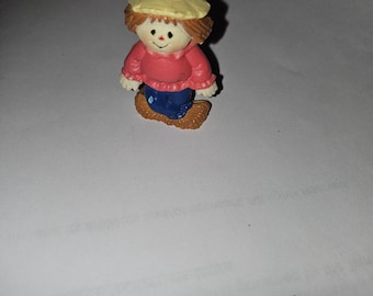 May include: A small scarecrow figurine. It features a light yellow hat, a red top, blue trousers, and brown boots. The figurine has a smiling face and is likely made of painted resin, suitable for display.