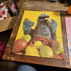 Puede incluir: Un personaje azul de Cookie Monster de Plaza Sésamo sosteniendo un plátano y una manzana. El rompecabezas presenta un fondo amarillo con una variedad de frutas, incluyendo manzanas, peras, uvas y plátanos.