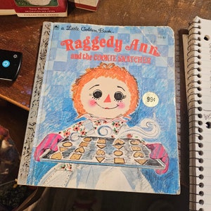 May include: A vintage Little Golden Book featuring Raggedy Ann and the Cookie Snatcher. The cover features a colorful illustration of Raggedy Ann holding a baking sheet of cookies. The book is blue and white with a checkered pattern and the title is in red and black.