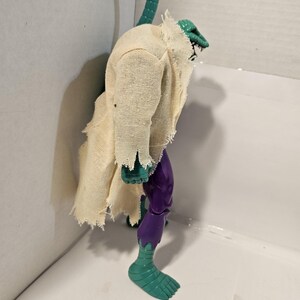 May include: A vintage action figure of a reptilian humanoid creature. The figure has a green head and tail, a tattered beige cloth garment, and purple and green limbs. The figure is posed in a standing position.