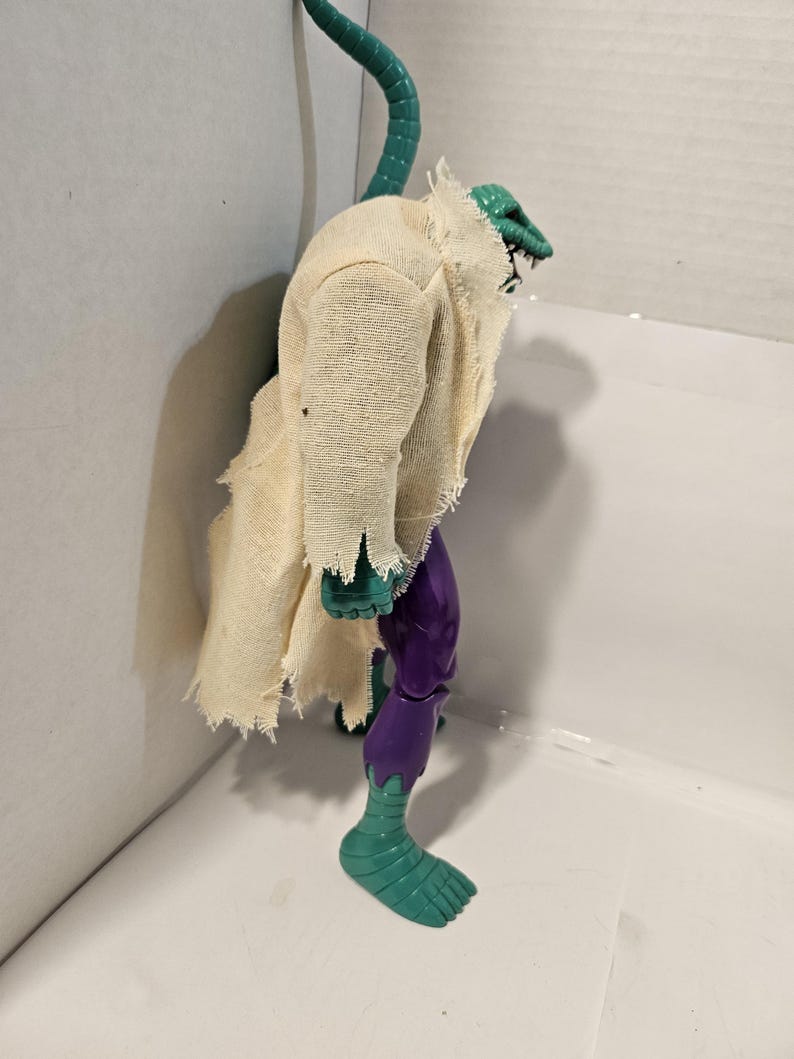 May include: A vintage action figure of a reptilian humanoid creature. The figure has a green head and tail, a tattered beige cloth garment, and purple and green limbs. The figure is posed in a standing position.