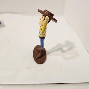 May include: A small Woody action figure from the Toy Story franchise. The figure is wearing a brown cowboy hat, a yellow shirt with a cow print vest, blue trousers, and brown boots. The figure stands on a brown oval base.