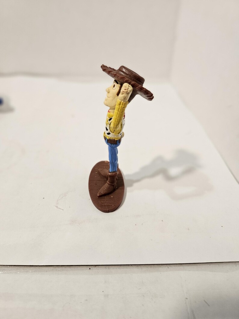 May include: A small Woody action figure from the Toy Story franchise. The figure is wearing a brown cowboy hat, a yellow shirt with a cow print vest, blue trousers, and brown boots. The figure stands on a brown oval base.