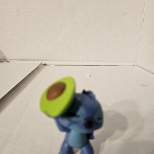 May include: A blue and teal Stitch figurine from Lilo & Stitch, holding a green and brown avocado slice. The figurine is standing on a white surface.