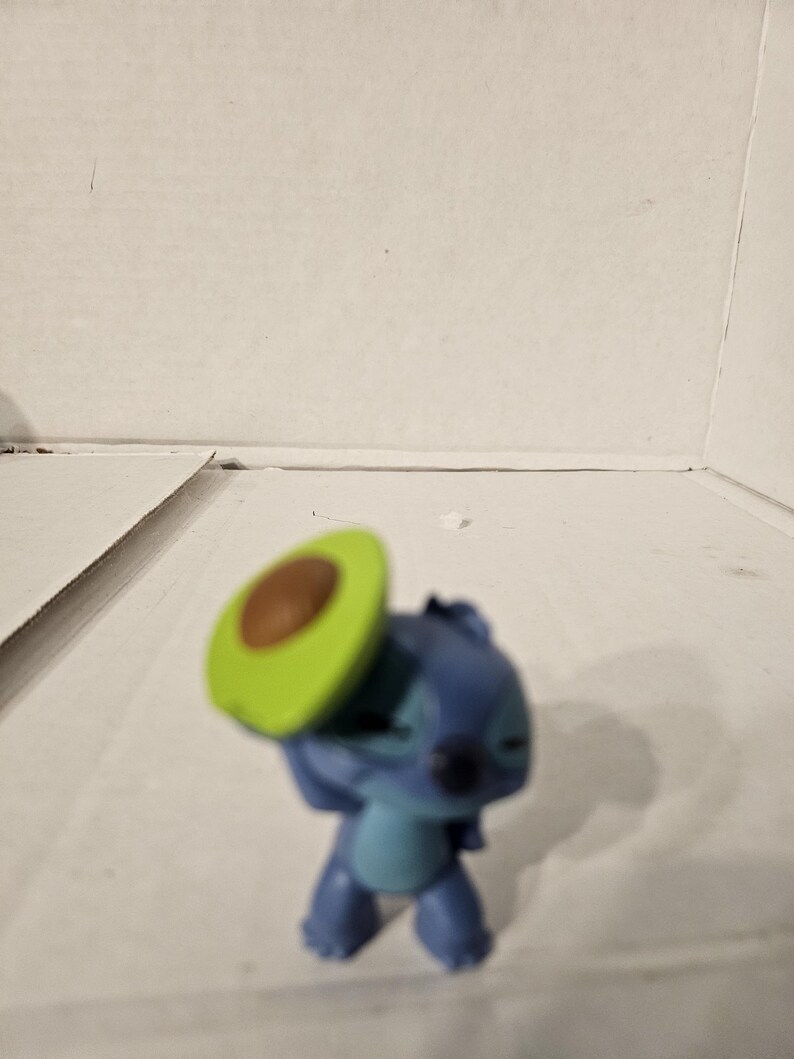 May include: A blue and teal Stitch figurine from Lilo & Stitch, holding a green and brown avocado slice. The figurine is standing on a white surface.