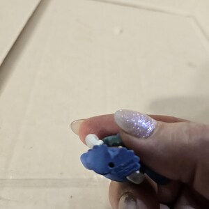 Pu&ograve; includere: Piccola statuetta blu con dettagli bianchi e verdi. La statuetta &egrave; tenuta in mano, con lo sfondo sfocato.