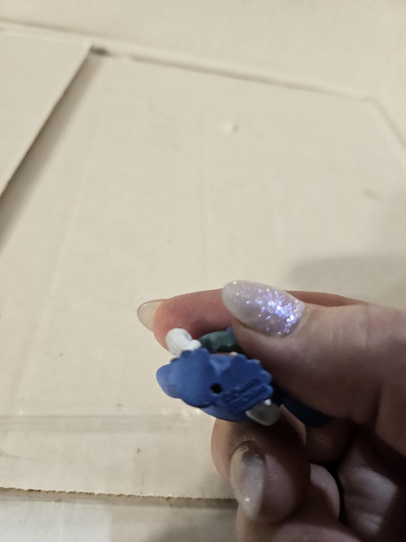 Pu&ograve; includere: Piccola statuetta blu con dettagli bianchi e verdi. La statuetta &egrave; tenuta in mano, con lo sfondo sfocato.