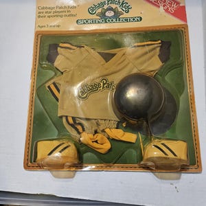May include: A vintage Cabbage Patch Kids Sporting Collection doll outfit, still in its original packaging. The outfit includes a tan jersey, shorts, hat, and shoes with black and yellow accents. The packaging reads "Six New Designs."